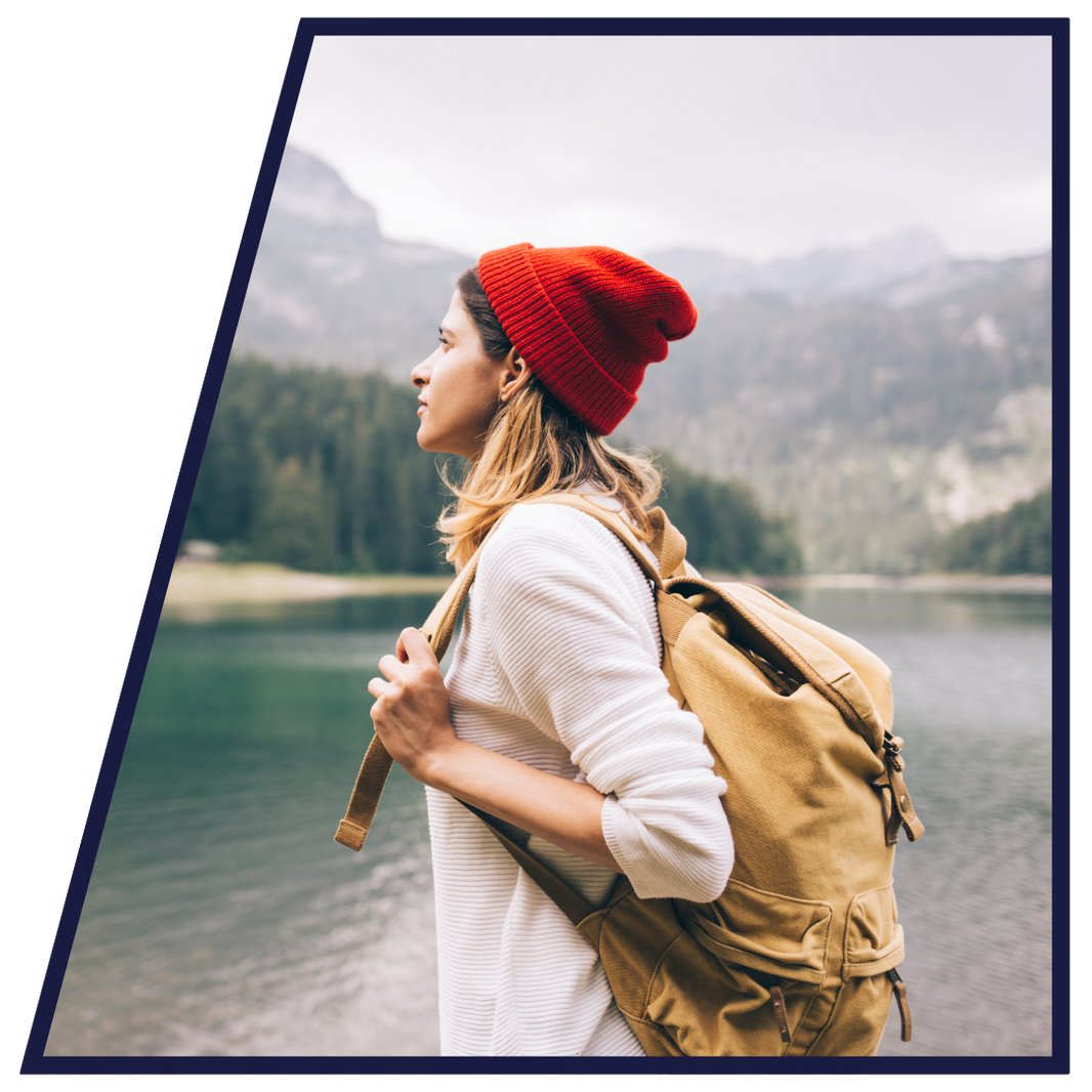 Woman with a red beanie and tan backpack standing by a lake with mountains in the background